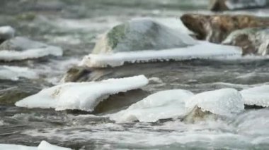 Kış nehri kar ve buzla kaplı taşların yakınından akıyor.