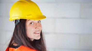 Sarı kasketli, turuncu renkli yüksek görüş yeleği olan, uzun siyah saçlı, omzunun üzerinden bakan, kendinden emin gülümseyen genç bir kadın. Bulanık inşaat alanı arka planı, metin sağ tarafı için boşluk