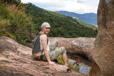 Yürüyüş elbiseli genç bir kadın, doğal su havuzu yakınlarındaki taşların üzerinde dinleniyor, bulanık ağaçlar ve dağların arka planında - Andringitra, Madagaskar 'daki yürüyüş sırasında görülen tipik manzara