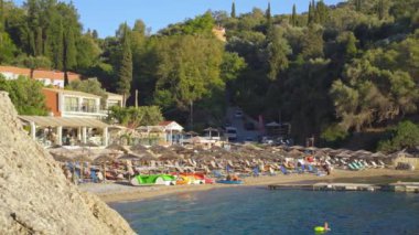 Liapades, Yunanistan - 21 Eylül 2022: Güneşli yaz gününde Liapades Körfezi plajında dinlenen insanlar, küçük dalgalarla denizde yüzen bir kişi
