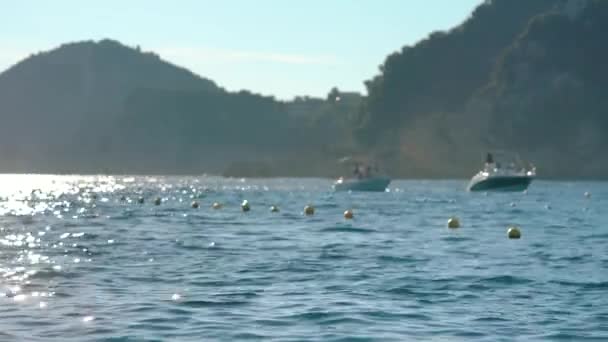 ギリシャのルピアデス湾の景色は 前景に小さな波で穏やかな海に焦点を当て 岩の崖の背景でゆっくりと移動するぼやけたボート アフタヌーンサンライトヘイズシーン — ストック動画