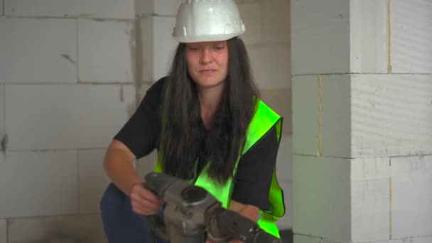 緑の高い可視性のベストおよび白い堅い帽子の若い女性の構造労働者は地面に電力ハンマー用具を置き 見回し 端に微笑みます — ストック動画