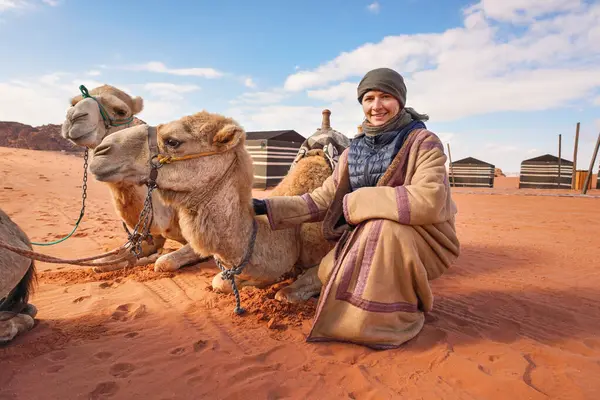 Young woman in traditional Bedouin coat - bisht - and headscarf crouching next to two camels laying on red desert ground, smiling, camp tents in background