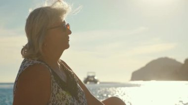 Gözlüklü, sakin deniz kenarında dinlenen yaşlı bir kadın, arka planda güneş ışığı, yakın çekim detayı.