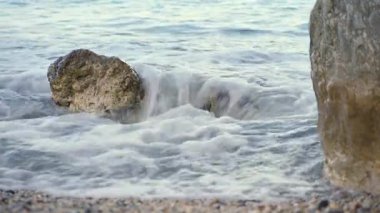 Kıyıdaki küçük deniz dalgaları kumlu sahildeki kayaları yıkıyor, sabahları yakın plan.