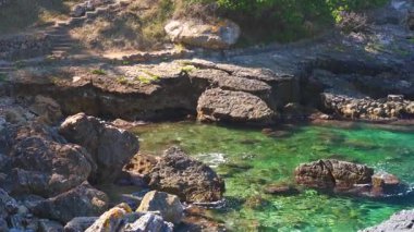 Kayalık sahil kıyısındaki oldukça sakin, berrak yeşil deniz, güneş küçük dalgalara parlıyor, kamera yavaşça yana dönüyor, Yunanistan 'ın Liapades kentindeki birçok koydan birinde tipik manzara