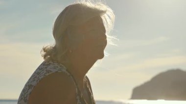 Gözlüklü, sakin deniz kenarında dinlenen yaşlı bir kadın, arka planda güneş ışığı, yakın çekim detayı.