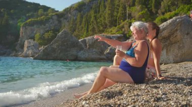 Gri saçlı yaşlı bir kadın, güneşli çakıl taşlı sahilde oturuyor, plastik bardakta içiyor, genç olanla konuşuyor, kızıyla konuşuyor, denizde bir şey gösteriyor ve işaret ediyor.