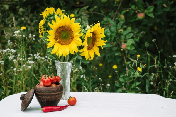 a bouquet of sunflowers on a white table with an autumn harvest of tomatoes and chili peppers