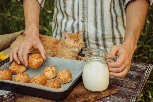 freshly baked pies in the hands of a man, buns on a baking sheet with milk on a brown wooden background, a red-haired cat near the baker