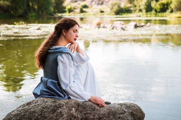 Uzun saçlı, tarihi mavi elbiseli bir kadın nehir kenarında oturuyor.