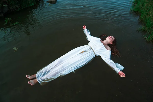 stock image a young woman in a white dress lies on the surface of the water in the river, medieval rituals 1