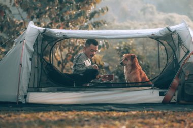 Adam sabahları kahve içer ve köpekle birlikte tatil için ormana kamp yaparken dinlenir. Meslek ve seyahat kavramı.