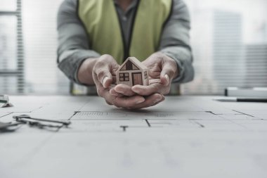Mimar mühendis elleri taslak üzerinde mini ev modelini tutuyor ve koruyor. Evi sat, koru, maliyet planlama ve inşaat konsepti.