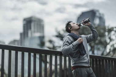 Genç sporcu adam koşudan sonra su içer, parkta koşar. Spor susuzluğu ve egzersizden sonra dinlenme. Büyük şehir yaşam konseptinde sağlık ve yaşam tarzı.