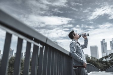 Genç sporcu adam koşudan sonra su içer, parkta koşar. Spor susuzluğu ve egzersizden sonra dinlenme. Büyük şehir yaşam konseptinde sağlık ve yaşam tarzı.