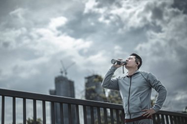 Genç sporcu adam koşudan sonra su içer, parkta koşar. Spor susuzluğu ve egzersizden sonra dinlenme. Büyük şehir yaşam konseptinde sağlık ve yaşam tarzı.