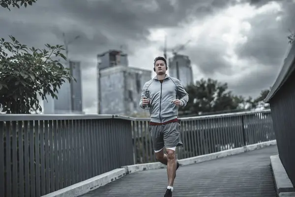 stock image A man holding a water bottle and running up on footbridge in the city center park for cardio workout.  Health and Lifestyle in big city life concept.