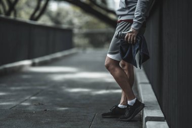  Yorgun adam dinleniyor ve nefes alıyor. Parktaki köprüde bir şişe içme suyu ve havlu tutuyor. Büyük şehir yaşam konseptinde sağlık ve yaşam tarzı.