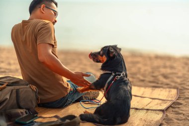 Bir adamın sahil kenarındaki bir paspasda oturan ve köpeğiyle oynayan bir köpeği var. Hayvan aile kavramı.