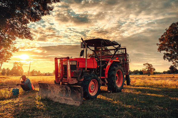 Трактор и прицеп припаркованы в поле, так как поблизости работает рабочая сила. Транспорт для сбора урожая. Солнце садится, бросая теплый свет на ландшафт и подчеркивая красоту сельской жизни.
