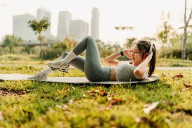 Spor kıyafetleri giymiş bir kadın kulaklık takıyor ve karın kaslarını çalıştırırken müzik dinliyor..