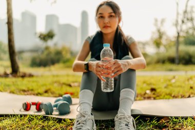 Spor kıyafetleri giymiş, susamış bir kadın, parkta egzersiz yaparken su şişesini açıyor..