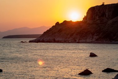 Arka planda kayalar ve dağlarla denizin üzerinde gün batımı. Sahilde gün batımı. Denizden yansıyan güneş ışığı.