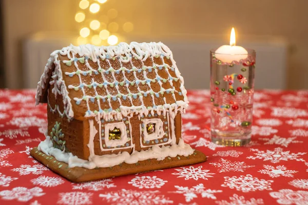Little gingerbread house with glaze standing on table with tablecloth and decorations, candles and lanterns. Living room with lights. Holiday mood. High quality photo