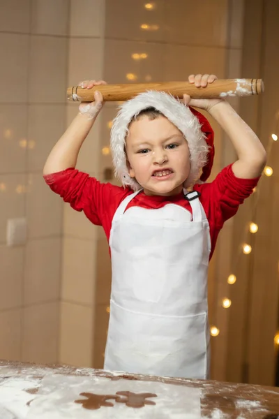 Evcil mutfağında oklavası olan komik çocuk. Çocuk Noel kurabiyesi pişiriyor. Çocuk evde Noel kurabiyesi hazırlıyor. Çocuk eğleniyor. Noel Baba şapkalı ve beyaz önlüklü çocuk kurabiye yapıyor. Yüksek