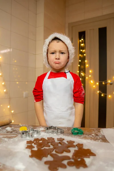Sad toddler child, standing under the table, while preparing Christmas cookies. Sad child in Santa hat. High quality photo