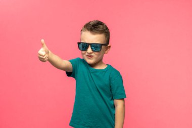 Pleasant satisfied boy shows thumbs up looking at camera, portrait. Attractive smiling boy in green casual t-shirt posing isolated on pink background. childhood, human emotions, lifestyle. High