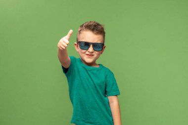 Cheerful little boy in sunglasses showing thumbs up gesture, isolated on green background. Copy space. High quality photo