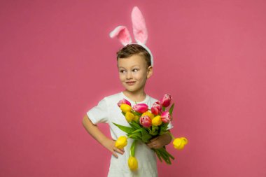 Beautiful cute little boy in Easter bunny ears smiling and holds spring bouquet of yellow and pink tulips on a pink background. High quality photo