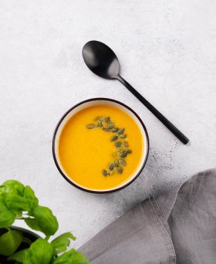 Sonbahar vejetaryen balkabağı çorbası kremalı ve tohumlu beyaz arka planda taze bitkisel ve kaşıklı. Sağlıklı yavaş yemek kavramı. Üst görünüm ve kopyalama alanı.