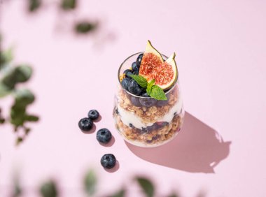 Tahıllı lezzetli yoğurt, incir ve yaban mersinleri pembe arka planda, yapraklı ve gölgeli bir bardakta. Sağlıklı ve besleyici bir kahvaltı. Diyet yemek. Üst görünüm ve kopyalama alanı.