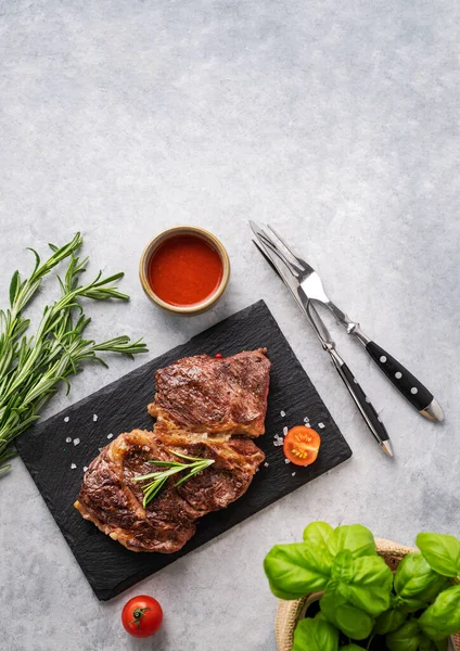 BBQ grilled beef steak with sauce, rosemary and basil on black slate on light  background. Top  view and copy space.