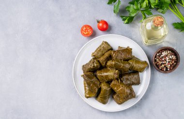 Dolma. Hafif arka planda zeytinyağı, domates ve biberli taze otlarla dolu mavi bir tabakta doldurulmuş üzüm yaprakları (geleneksel Kafkas, Türk, Yunan mutfağı). Üst görünüm ve kopyalama alanı.