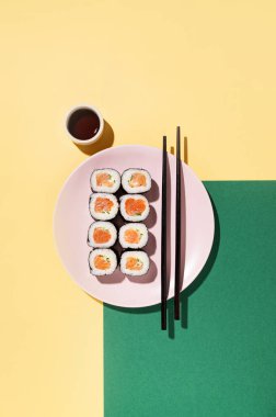 Japon suşisi, somon, salatalık ve soya sosuyla sarılı pembe tabakta yemek çubukları ve sert gölgeli yeşil arka planda. Üst görünüm ve kopyalama alanı.