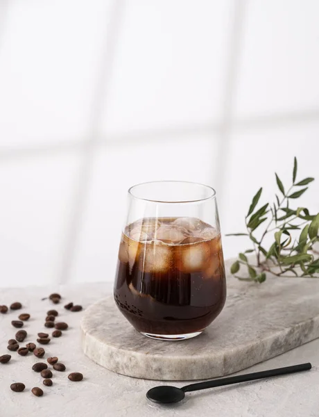 Iced coffee in a glass with ice cubes and grains on a light marble background with morning shadows. The concept of a cold summer drink. Copy space.