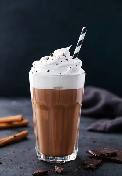 Hot chocolate with whipped cream, tube and cinnamon in a tall glass on a dark background. Front view and copy space.