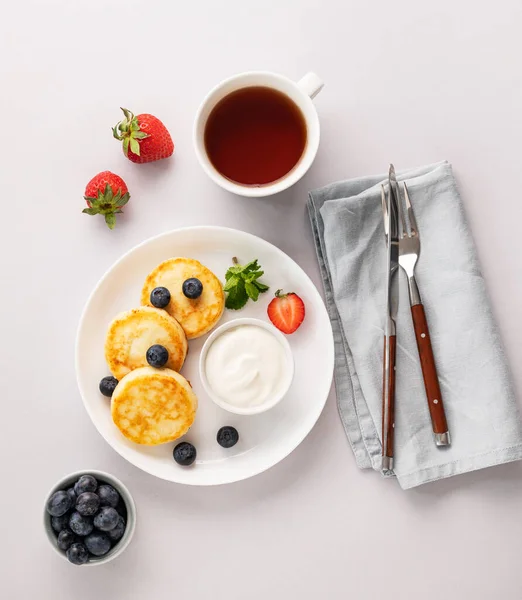 stock image Pancakes with cottage cheese with fresh blerries and sour cream on a gray background with a cup of tea. The concept of a healthy and delicious breakfast. Top view.