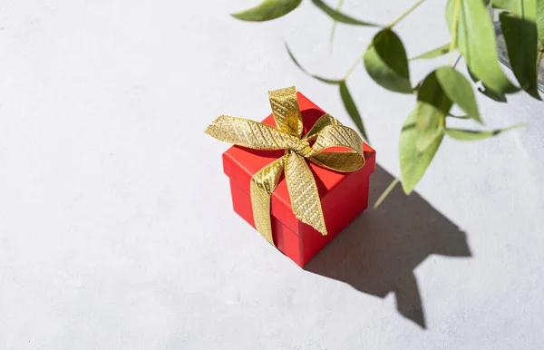 A red gift box with a gold ribbon on a gray background with shadow and branch flower. The concept of holiday photography. Surprise for Valentine\'s Day, birthday, wedding. Copy space and top view.
