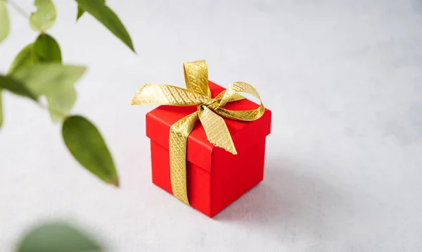 A red gift box with a gold ribbon on a gray background with branch flower. The concept of holiday photography. Surprise for Valentine\'s Day, birthday, wedding. Copy space and top view.