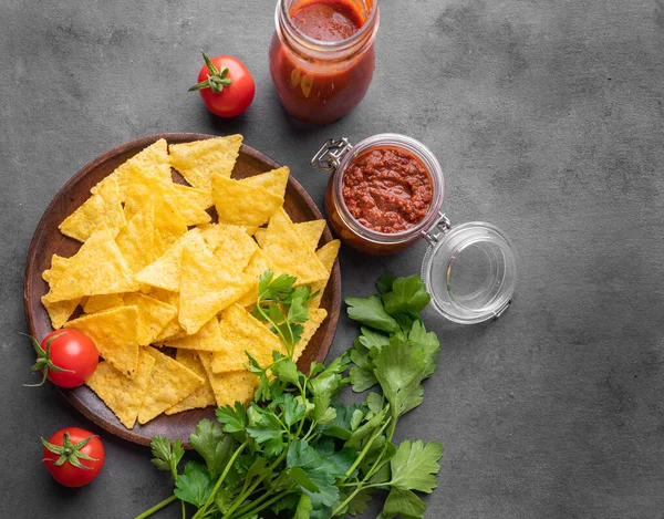 stock image Nachos or tortilla with spicy tomato sauce on a plate with fresh vegetables and herbs on a dark background. Corn chips with salsa sauce. Classic Mexican snack. Top view and copy space.