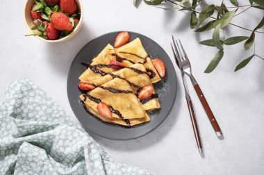 Taze çilekli tatlı krepler ve açık arkaplanda gri bir tabakta çikolata. Ev yapımı sağlıklı yiyecek kavramı. Metin için üst görünüm ve boşluk.