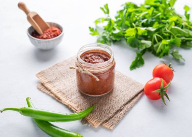 Hot sauce adjika. Homemade appetizer with pepper and tomatoes in a jar on light background with fresh herb and vegetables. The concept of Caucasian and Georgian food. clipart