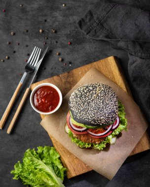 Sağlıklı vejetaryen burger, siyah çörek, nohut pirzolası, salata, salata, salatalık ve domates sosu ve koyu arkaplanda baharatlar. Vejetaryen fast food konsepti. Üst görünüm.