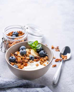 Vejetaryen yoğurt, muz ve yaban mersinli müsli, arka planda hafif bir kapta. Kahvaltı için sağlıklı ve besleyici ev yapımı tahıl gevreği. Metin için boş alan.