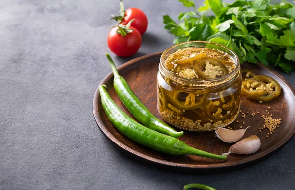 stock image Jalapeno hot pepper, sliced in a jar with spices on a wooden plate with fresh vegetables on a dark background. Vegetable vegetarian snack. Traditional Mexican cuisine.  Copy space.